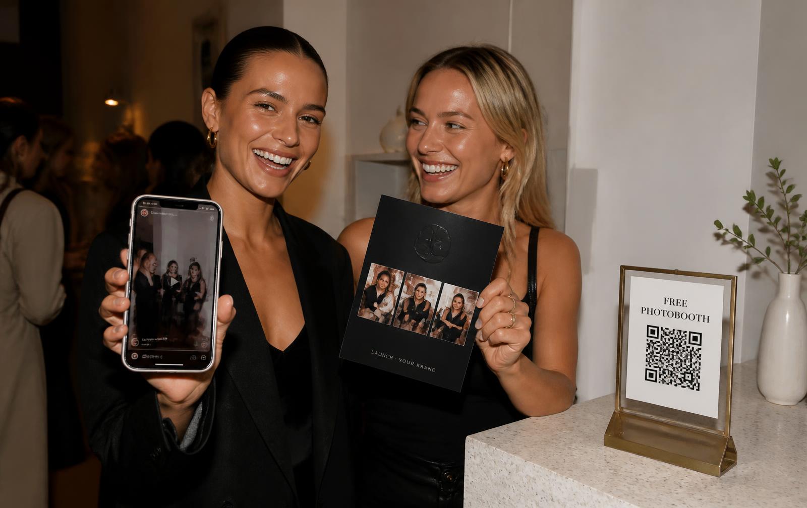 Two guests at a popup launch: one holds up her phone with the clip she just shared, the other a glossy black branded postcard, next to a brass “Free Photobooth” QR holder